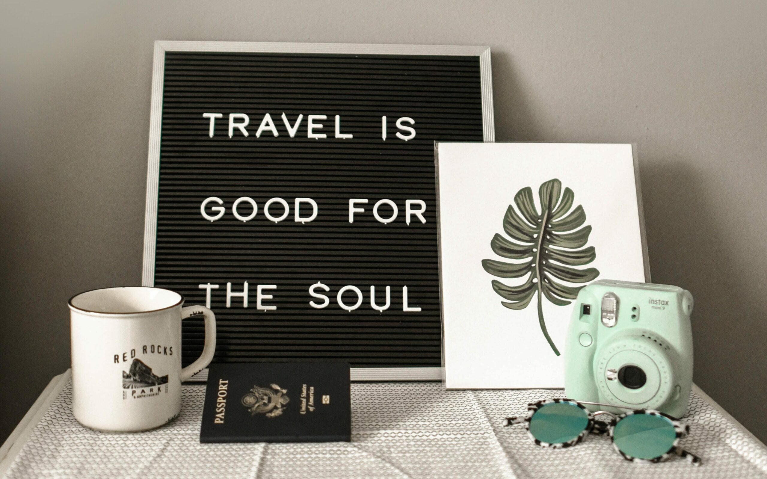 Napis "Travel is good for the soul", ilustracija monstere, polaroidni fotoaparat, skodelica, potni list in očala na mizi. 